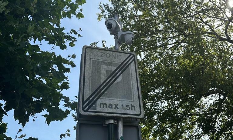 Camera boven een verkeersbord aan een paal, gericht op de straat, tegen bomen en lucht