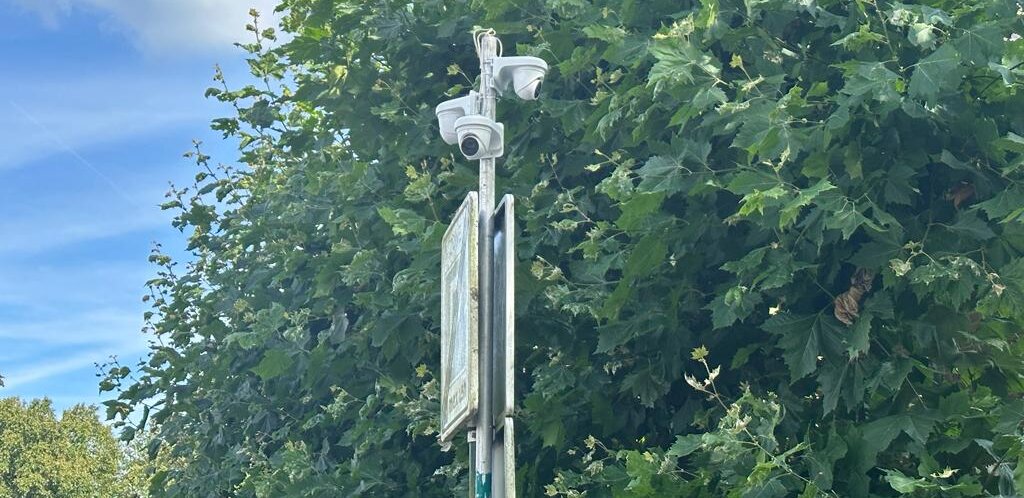 Camera's boven aan een paal naast een verkeersbord, met dichte groene bomen op de achtergrond