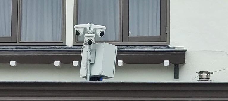 Twee witte camera's met een grijze systeemkast, gemonteerd tegen een gevel onder de dakgoot in de openbare ruimte