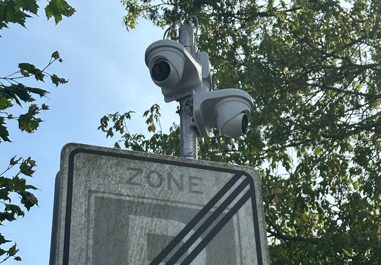 Twee witte camera's boven een verkeersbord aan een paal, tegen bomen en een blauwe lucht in de openbare ruimte