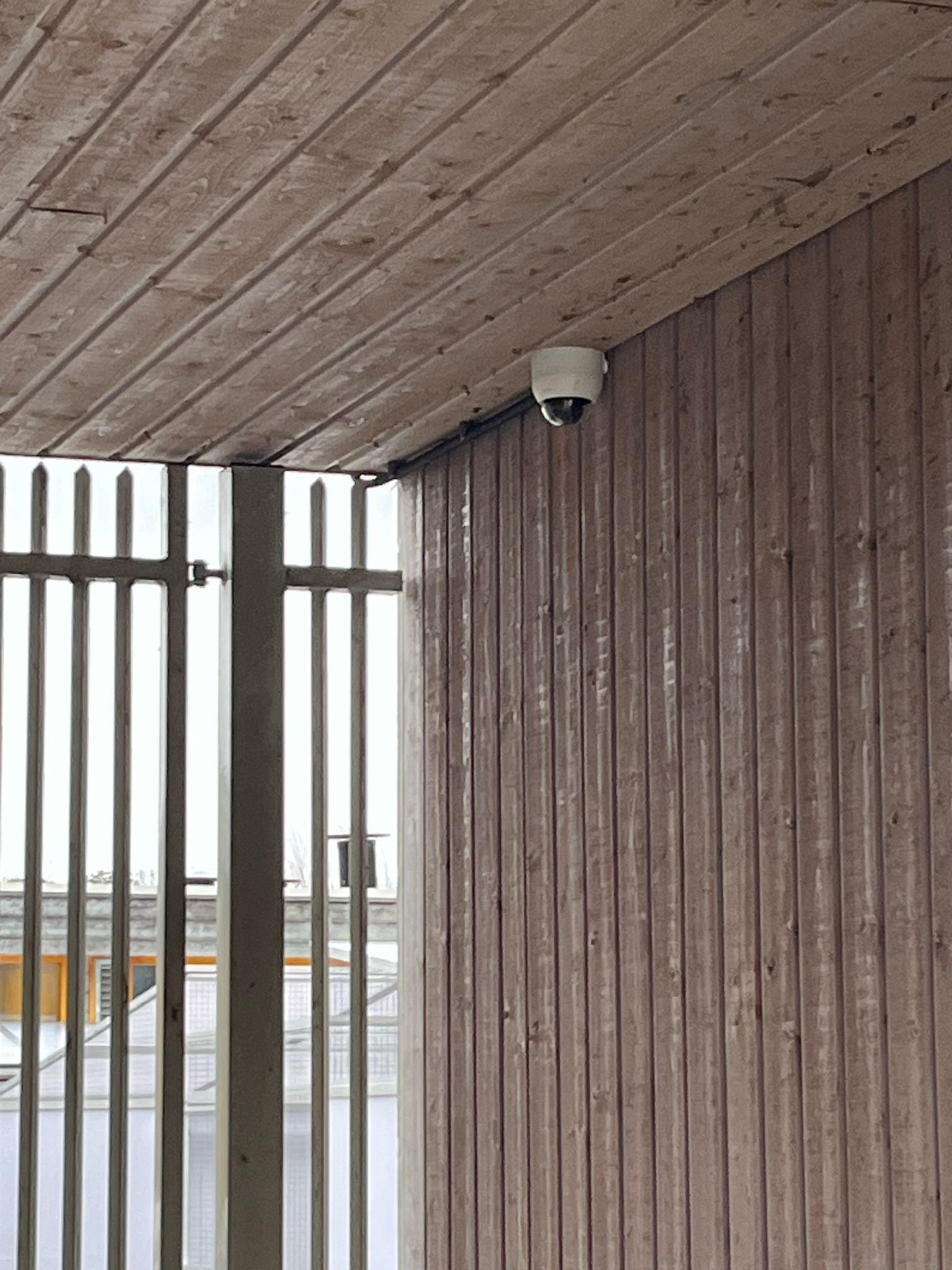 Witte domecamera onder een houten overkapping, naast een stalen hekwerk van een dierenverblijf