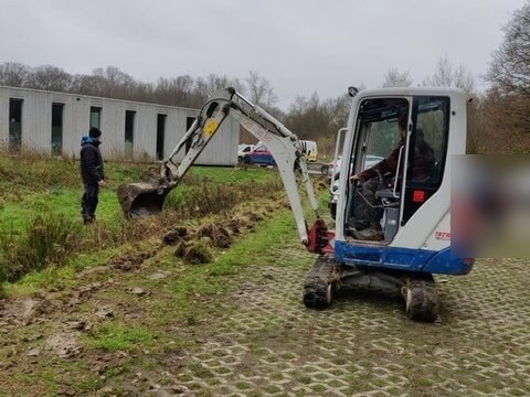 Minigraafmachine graaft een sleuf in de grasstrook langs een gebouw op het terrein, met een monteur ernaast
