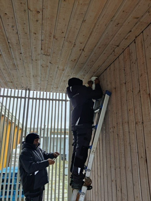 Twee monteurs van CameraInstallatie.nl monteren vanaf een ladder een camera tegen het houten plafond bij de dierenverblijven