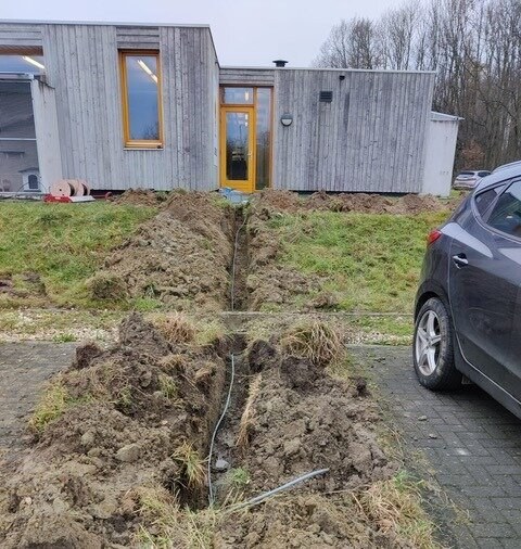 Open gegraven sleuven in het gras voor een houten gebouw, met een kabel in de sleuf