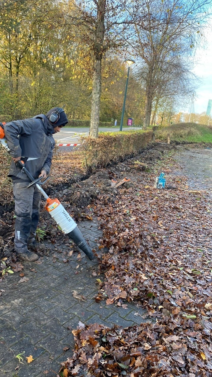 Collega blaast met een bladblazer het bestrating schoon langs de dichtgemaakte kabelsleuf naast de haag
