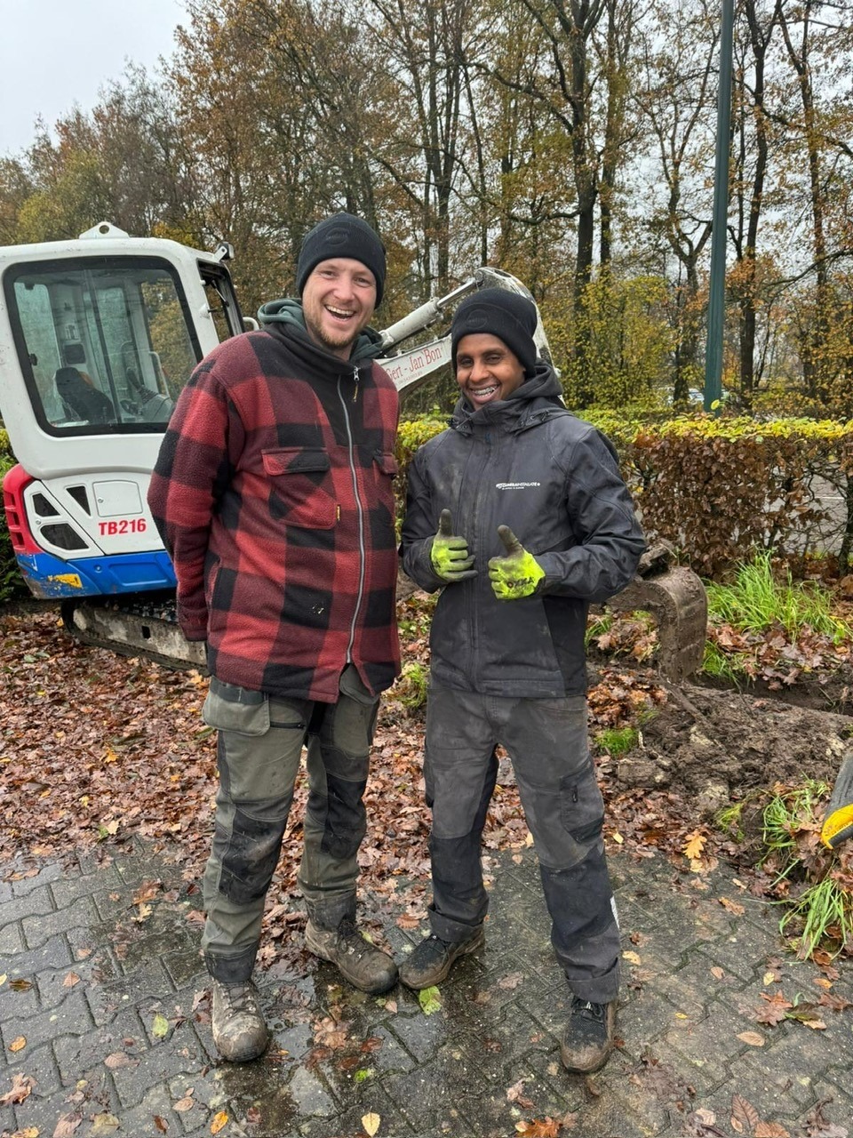 Twee collega&rsquo;s in werkkleding bij de minigraver op het parkterrein, na het graven van de kabelsleuf