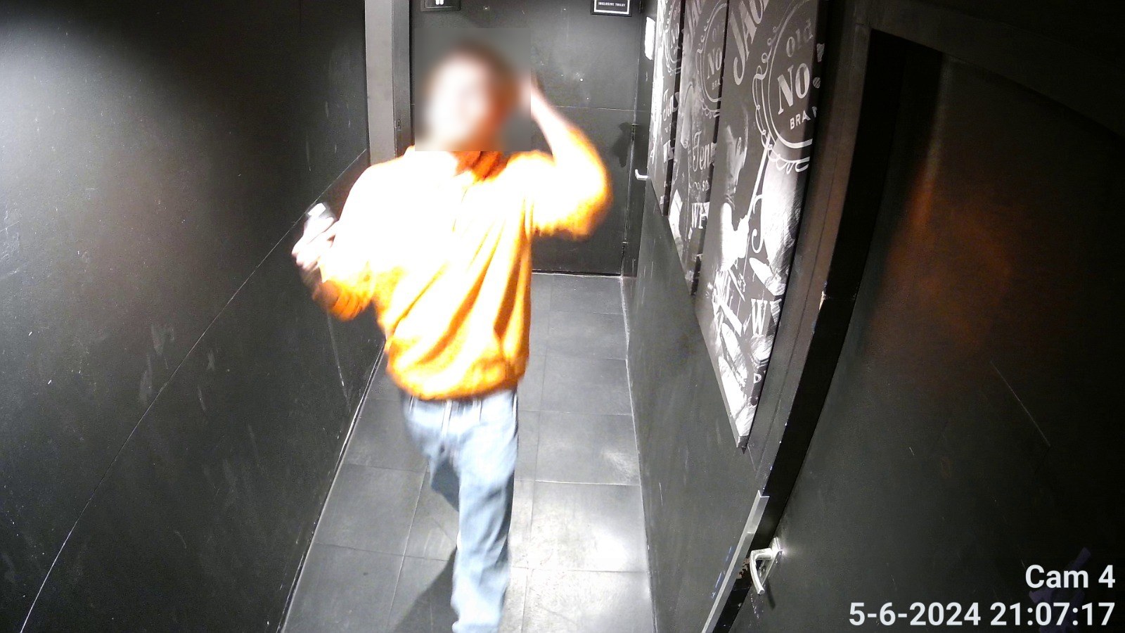 Camerabeeld van een smalle donkere gang waarin een persoon met een felverlichte trui loopt