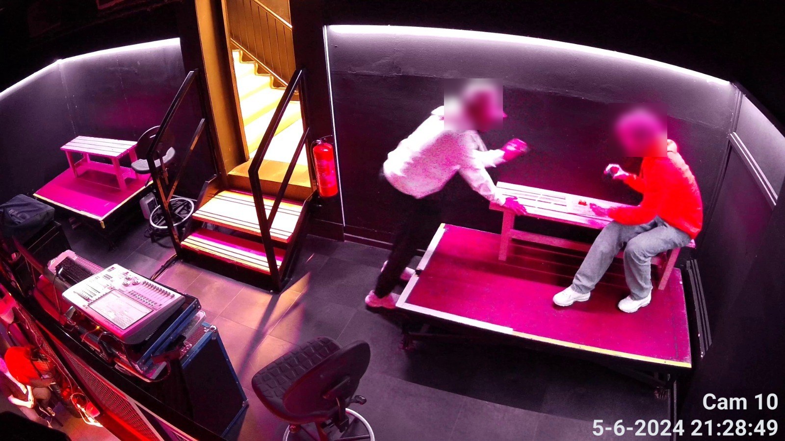 Camerabeeld van een kleine zaal met roze podiumverlichting, een geluidstafel links en twee personen aan een tafel