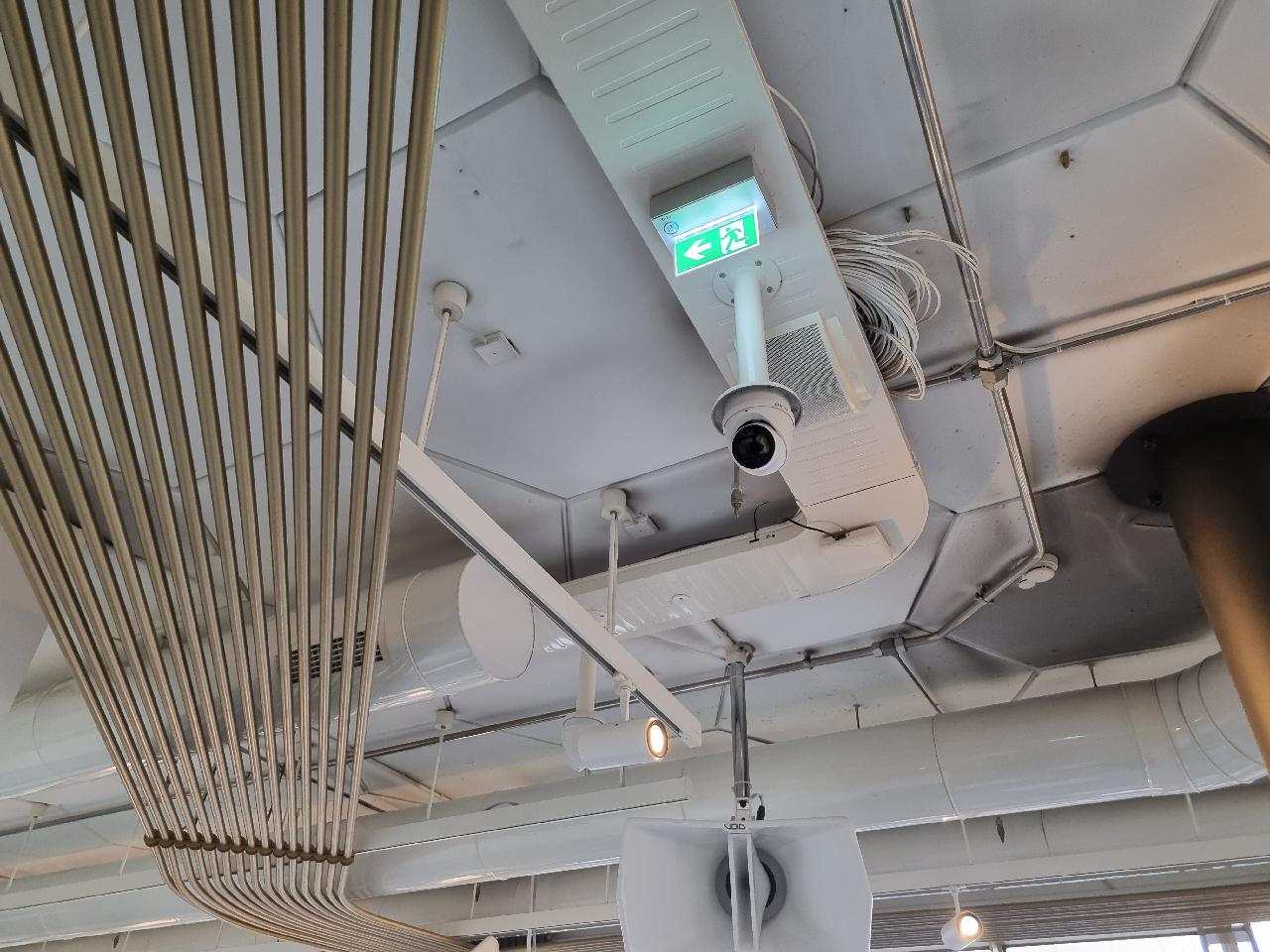 Witte dome-camera aan een technisch plafond, naast luchtkanalen en een opgerolde kabelbundel