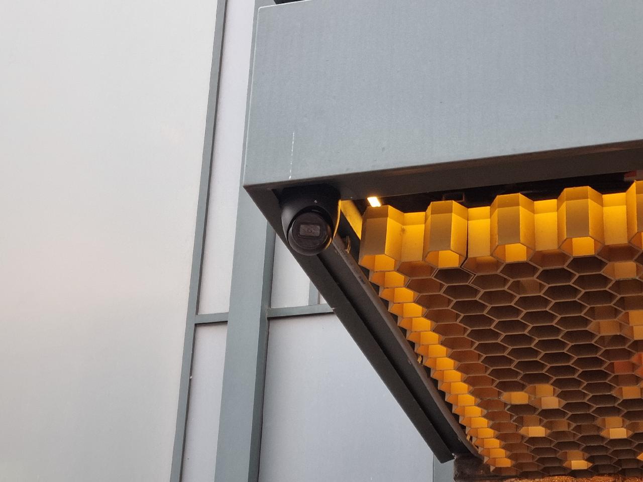 Zwarte camera gemonteerd onder de rand van een buitenoverkapping met verlichte gevelbekleding