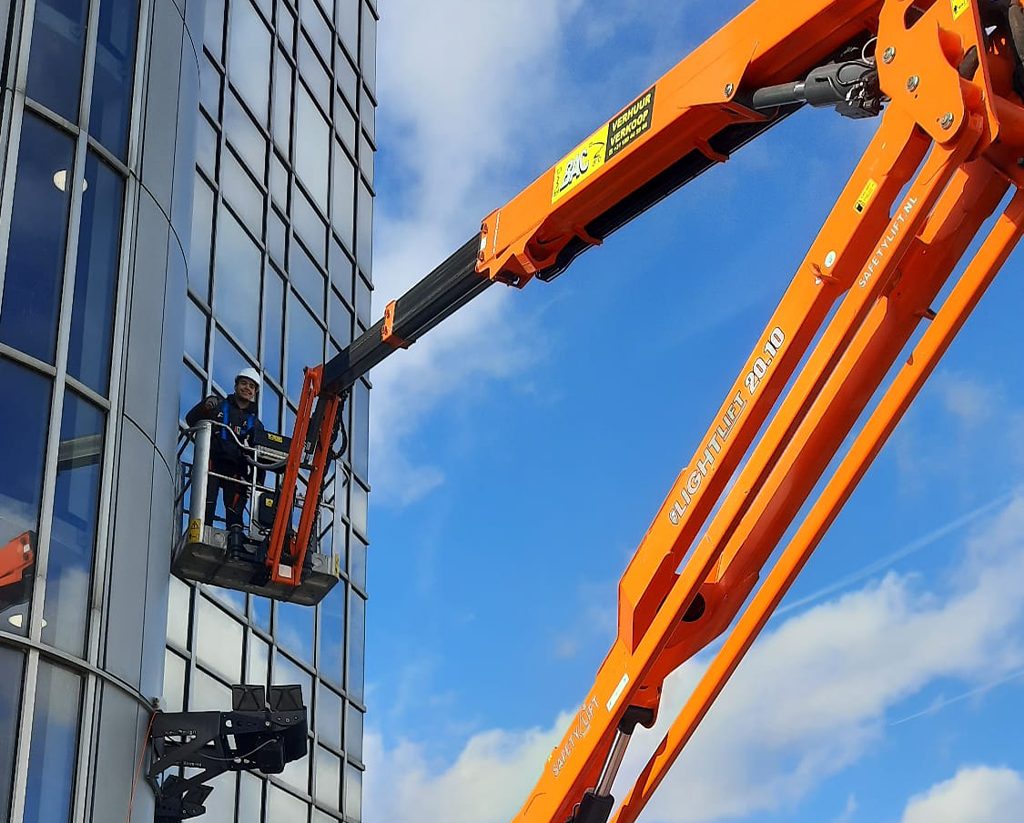 Monteur van CameraInstallatie met helm in de bak van een oranje spinhoogwerker langs een gebogen glazen toren
