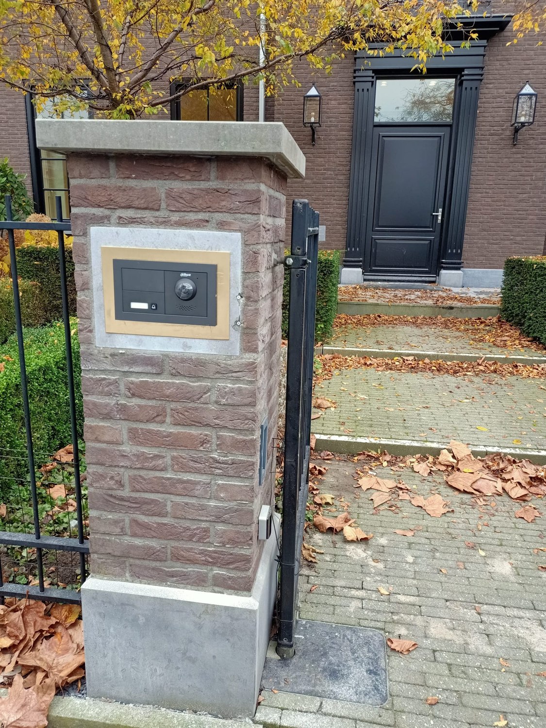 Video-intercom op een gemetselde poortpaal, met het hek open en het pad naar de voordeur erachter