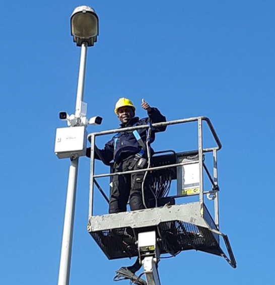 Monteur met helm in een hoogwerker steekt zijn duim op naast een lichtmast met armatuur tegen een blauwe lucht