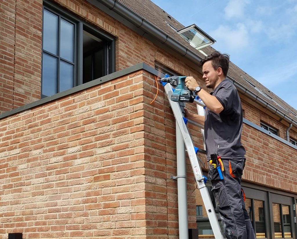 Onze monteur staat op een ladder tegen de gevel van een woning en boort een doorvoer voor de netwerkkabel naar het access point