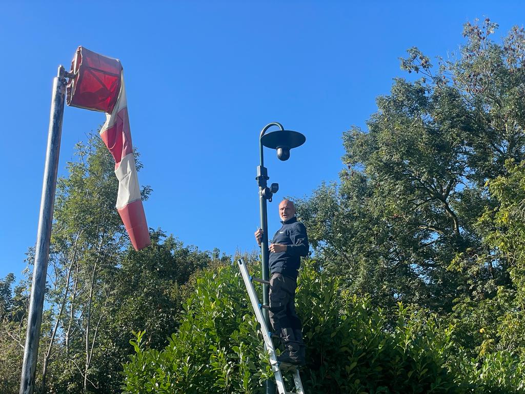 Monteur van CameraInstallatie op een ladder bij een camera op een paal, met bomen op de achtergrond