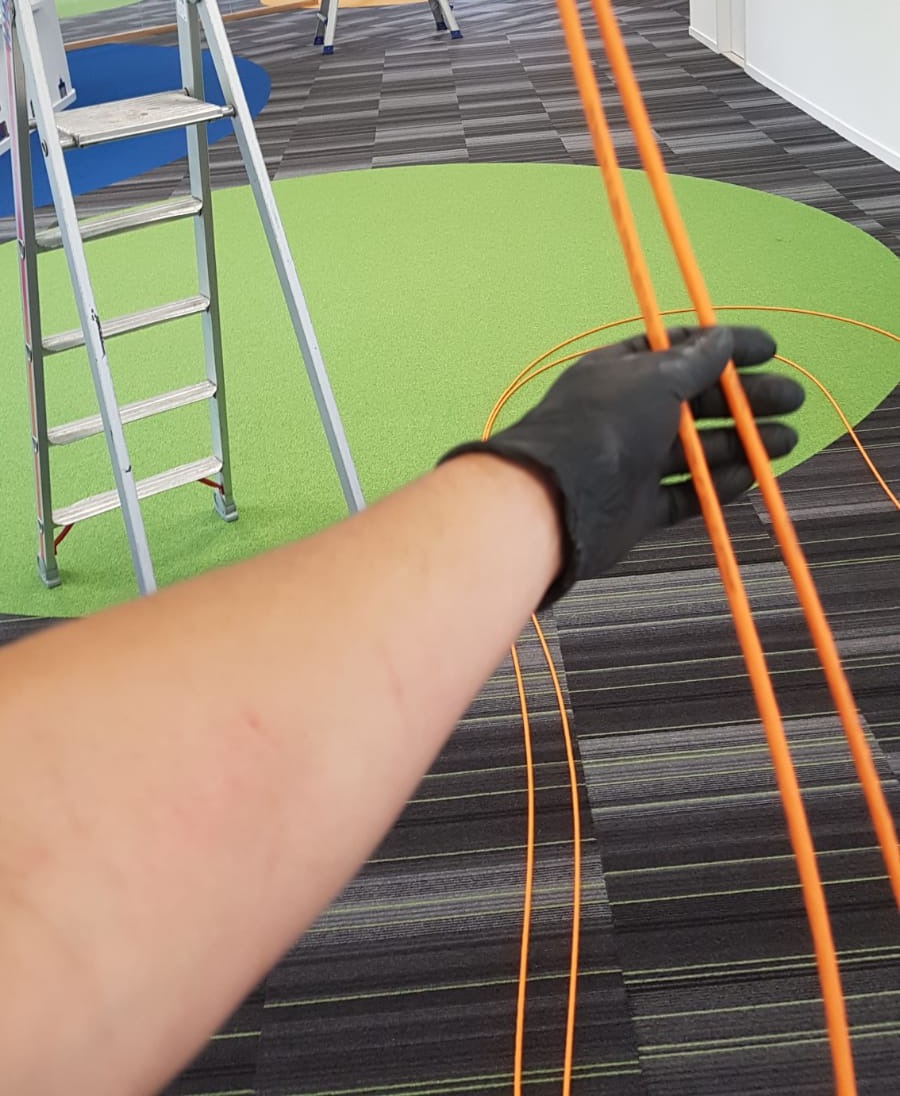 Hand met werkhandschoen trekt twee oranje netwerkkabels strak over het groene zitkleed van een leeshoek, met een ladder ernaast