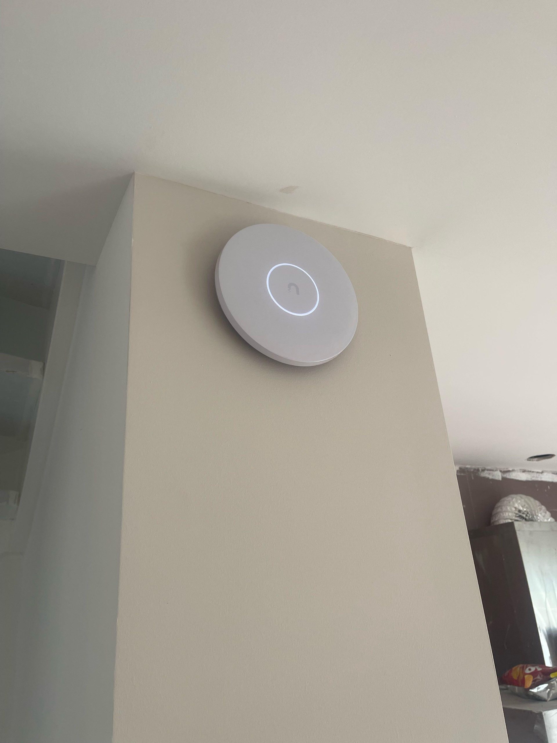 Wit rond UniFi wifi access point met verlichte ring, netjes tegen een binnenmuur bij het plafond gemonteerd