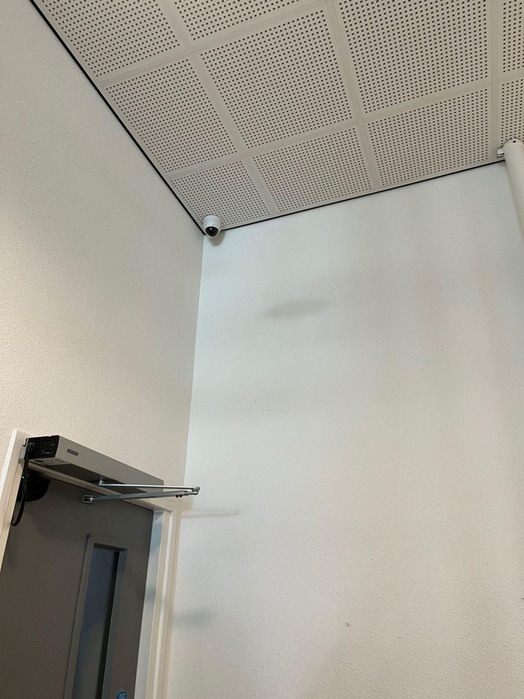 Witte dome-camera in de hoek tegen het systeemplafond van een gemeenschappelijke gang, boven een deur met deurdranger