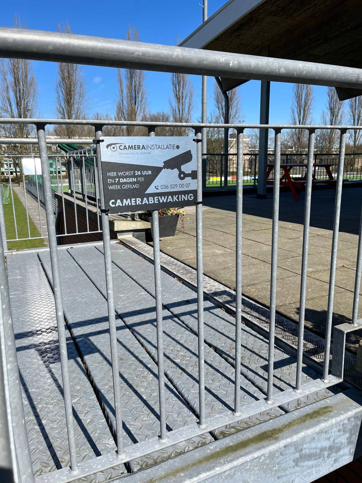 Waarschuwingsbord camerabewaking aan een hekwerk langs het terrein