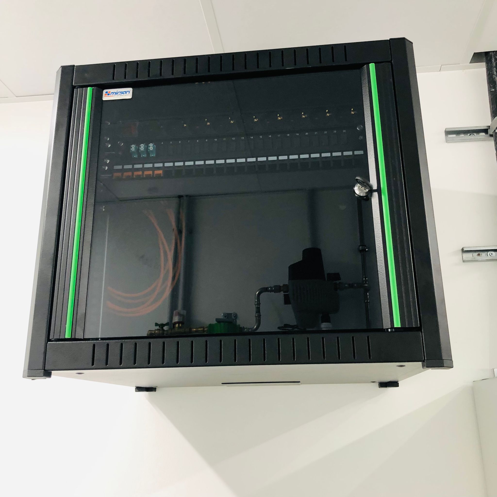 Zwarte wandpatchkast met netwerkapparatuur en groene statuslampjes achter een glazen deur