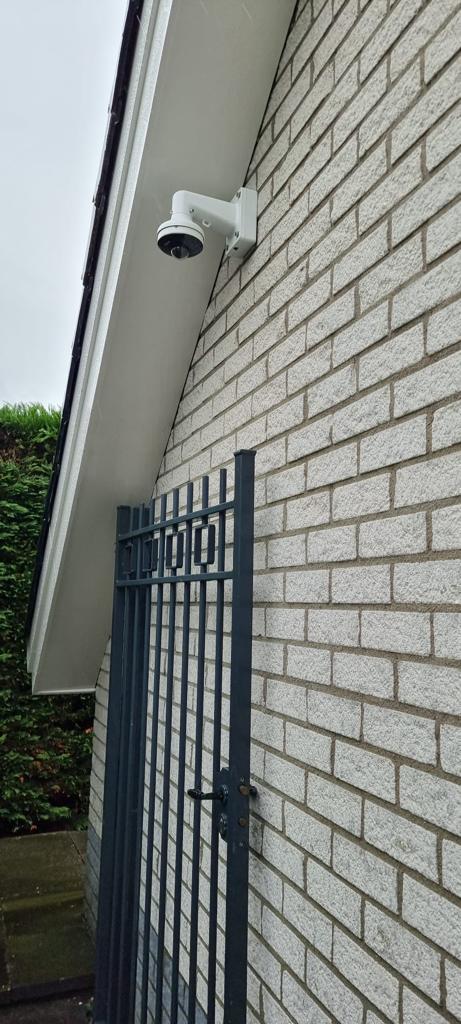Witte dome-camera op een beugel onder de dakrand aan de zijgevel van een woning, met zicht op de poort