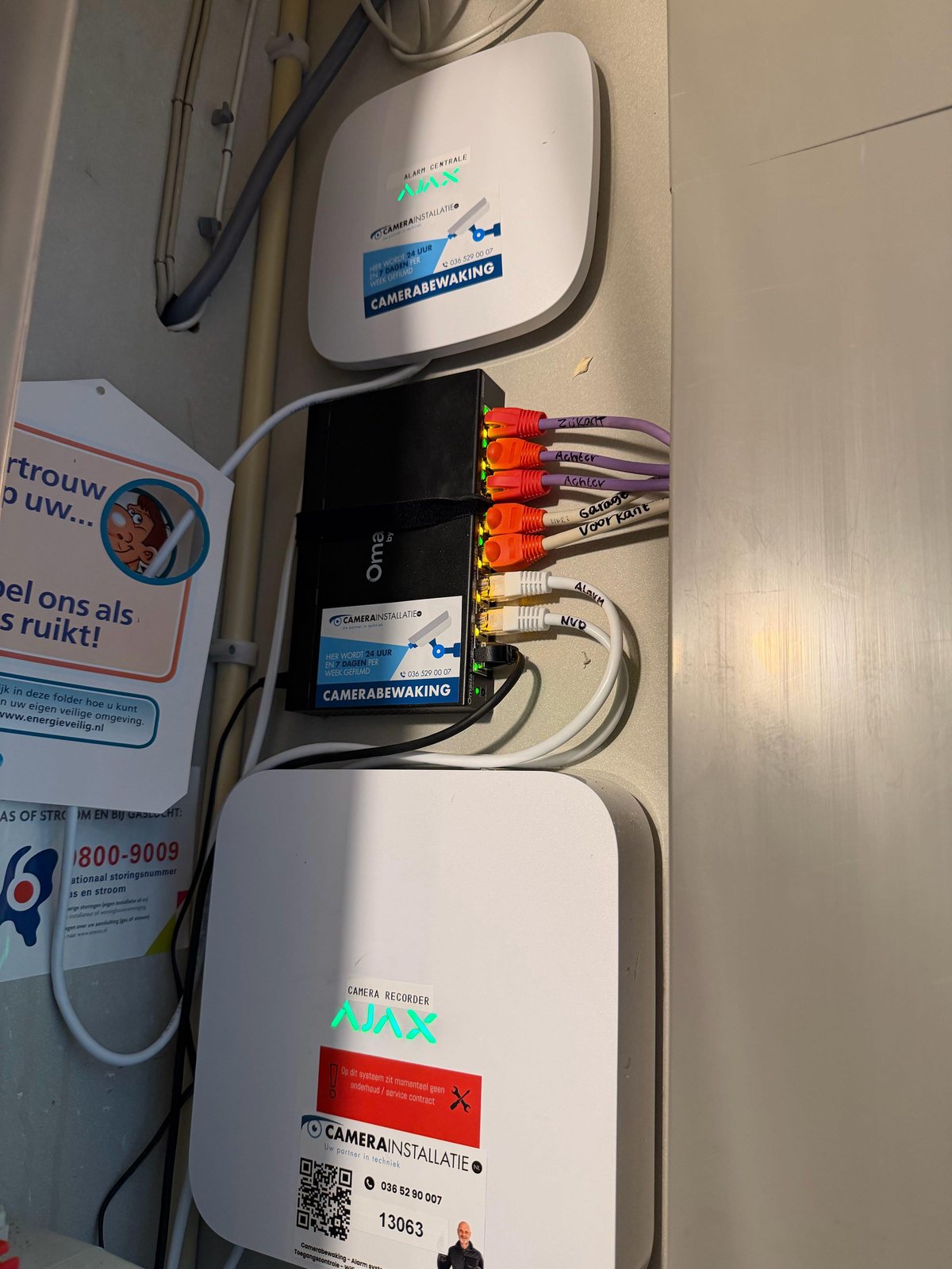 Alarmhub en recorder onder elkaar in de meterkast, met genummerde oranje patchkabels ertussen