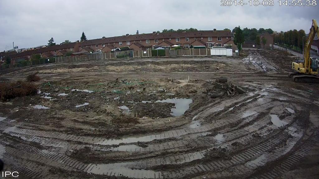 Camerabeeld van het draadloze systeem: breed overzicht van een open terrein met een graafmachine en een rij woningen op de achtergrond