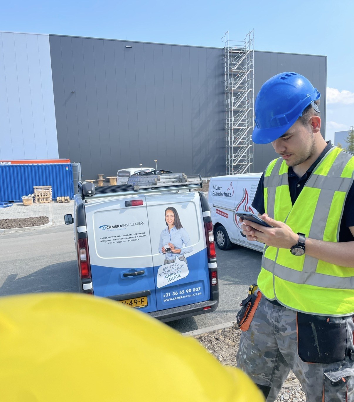 Monteur met helm en veiligheidshesje controleert de tekening op een tablet, naast onze bus op de nieuwbouwlocatie