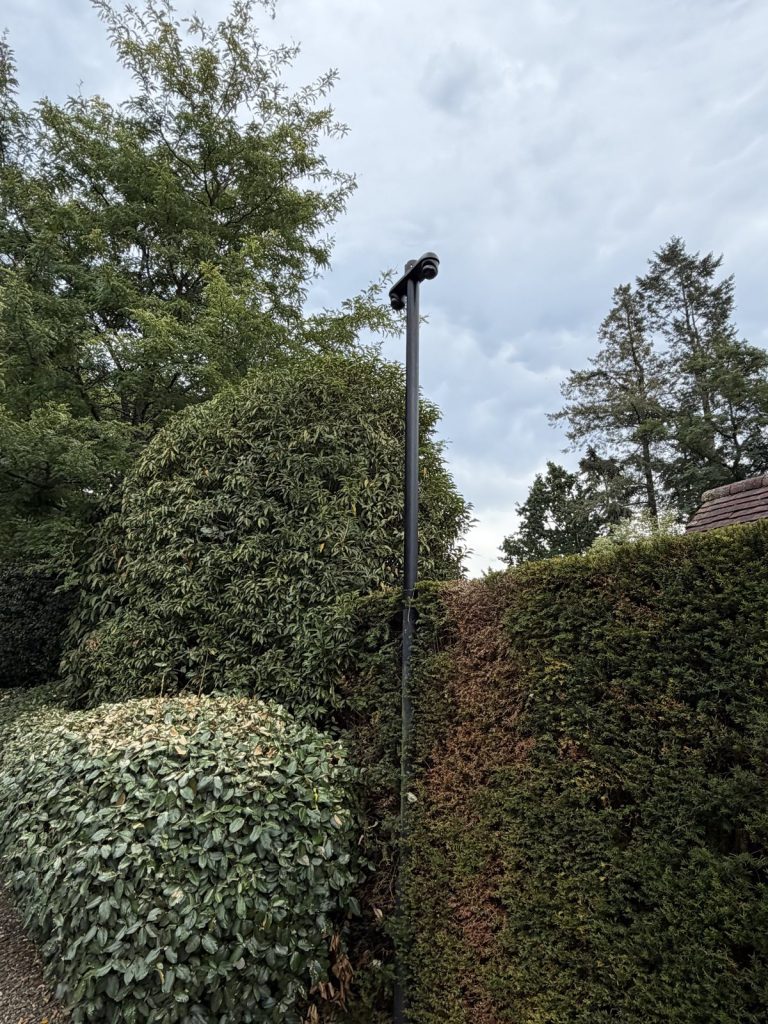 Zwarte cameramast in de achtertuin, weggewerkt tegen een hoge haag in Blaricum
