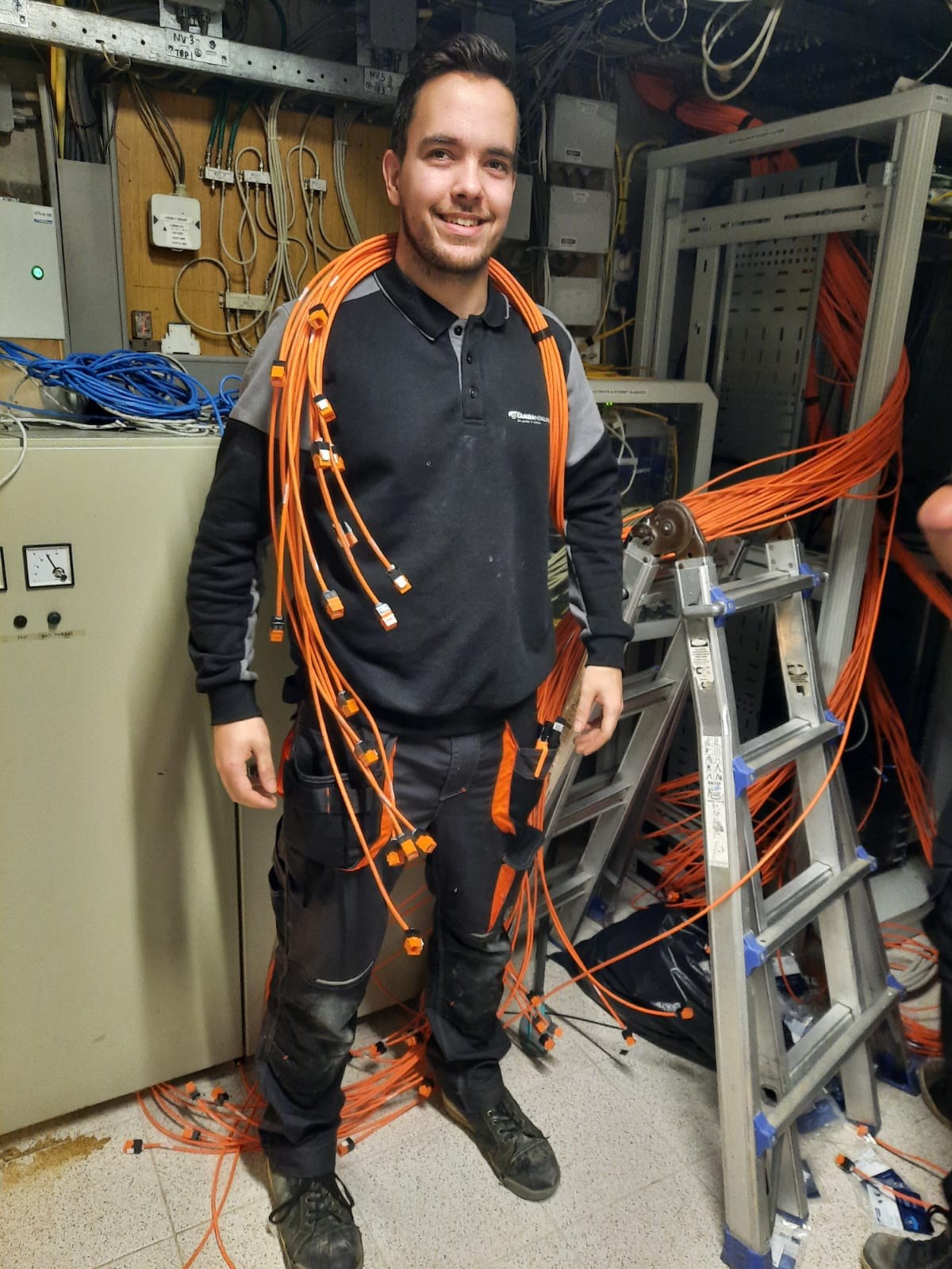 Lachende monteur van CameraInstallatie met een bundel oranje netwerkkabels om zijn nek in een technische ruimte