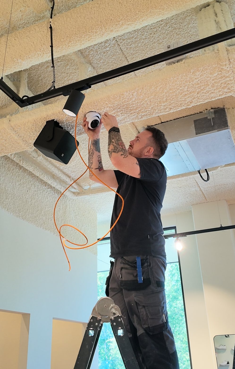Monteur van CameraInstallatie monteert vanaf een trap een dome-camera aan het plafond, met de oranje netwerkkabel al getrokken