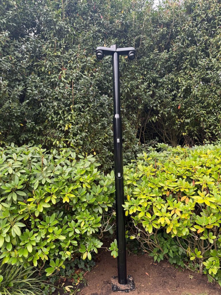 Zwarte cameramast die tegen een hoge groene haag vrijwel wegvalt