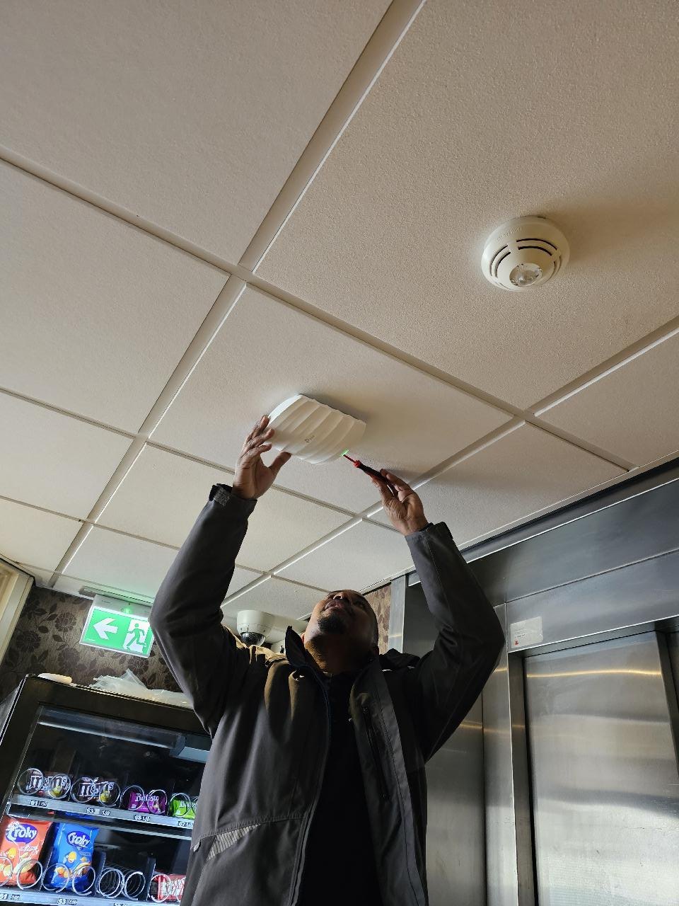 Monteur bevestigt een wit access point tussen de plafondplaten, naast een rookmelder