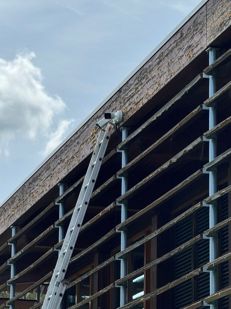 Monteur monteert een beveiligingscamera vanaf een ladder onder de dakrand van een bedrijfspand