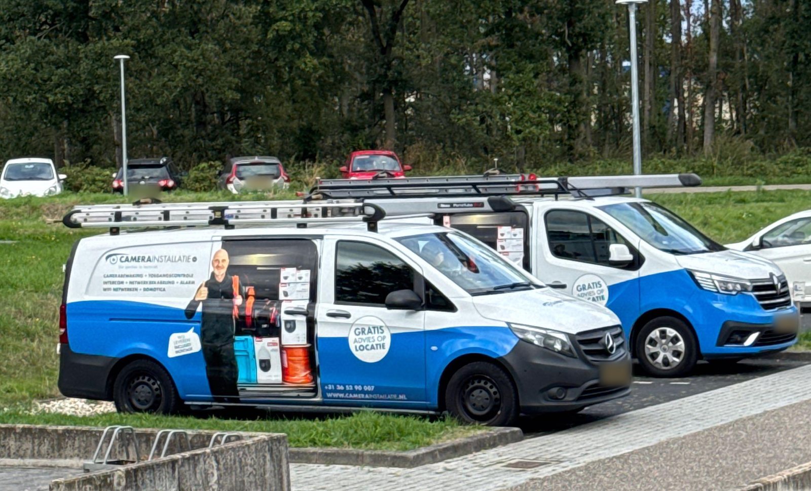 Twee bedrijfsbussen van CameraInstallatie.nl met ladders op het dak, geparkeerd op het terrein van de opvanglocatie