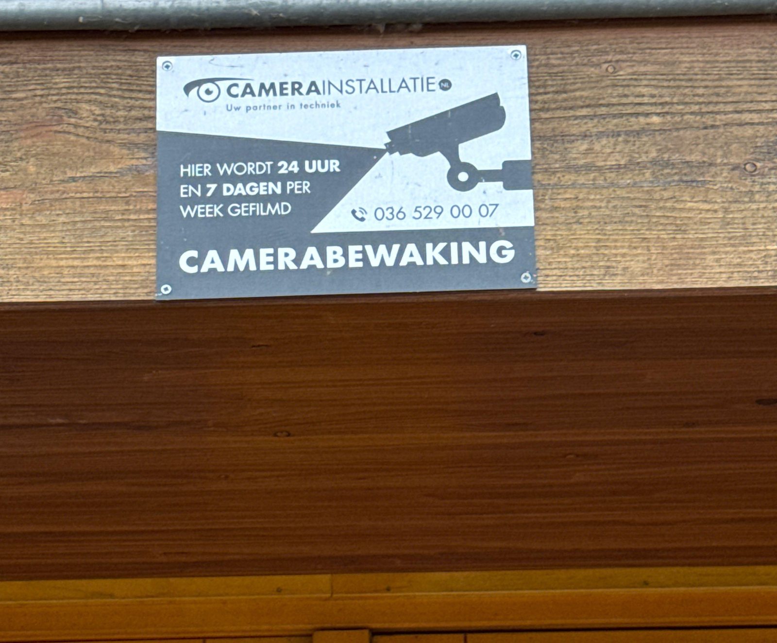 Metalen bord camerabewaking van CameraInstallatie.nl op een houten balk boven een deur
