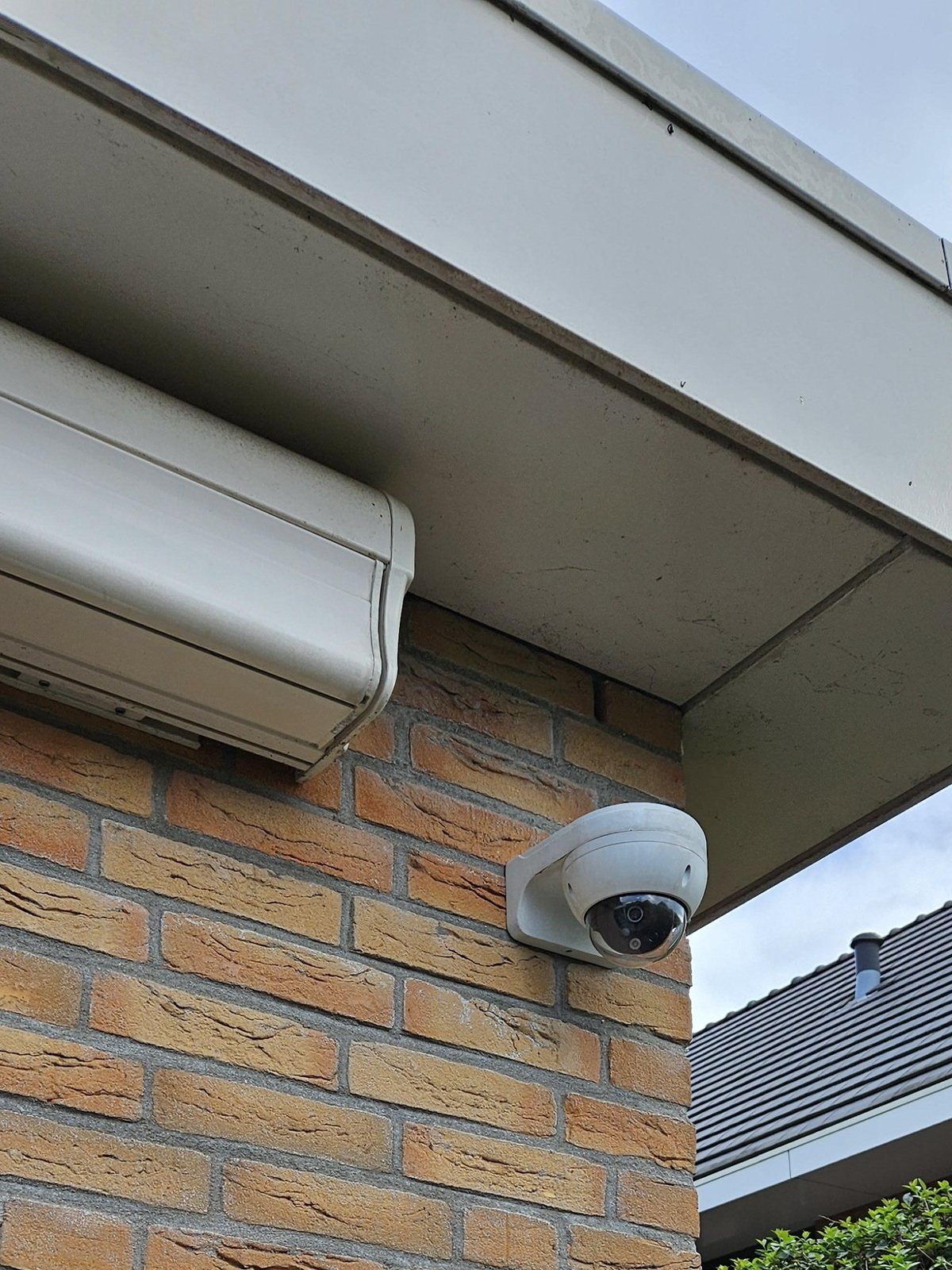 Witte dome-camera op een wandsteun, op de hoek van de gevel onder de dakrand, naast de kast van het zonnescherm