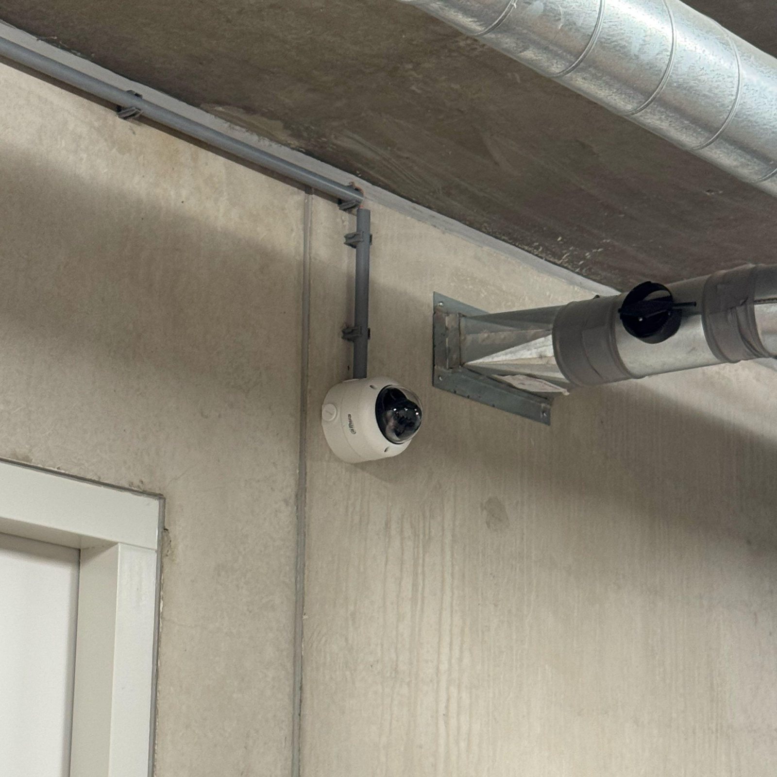 Witte Dahua-domecamera op een betonwand naast een deur, met de kabel in grijze buis langs het plafond