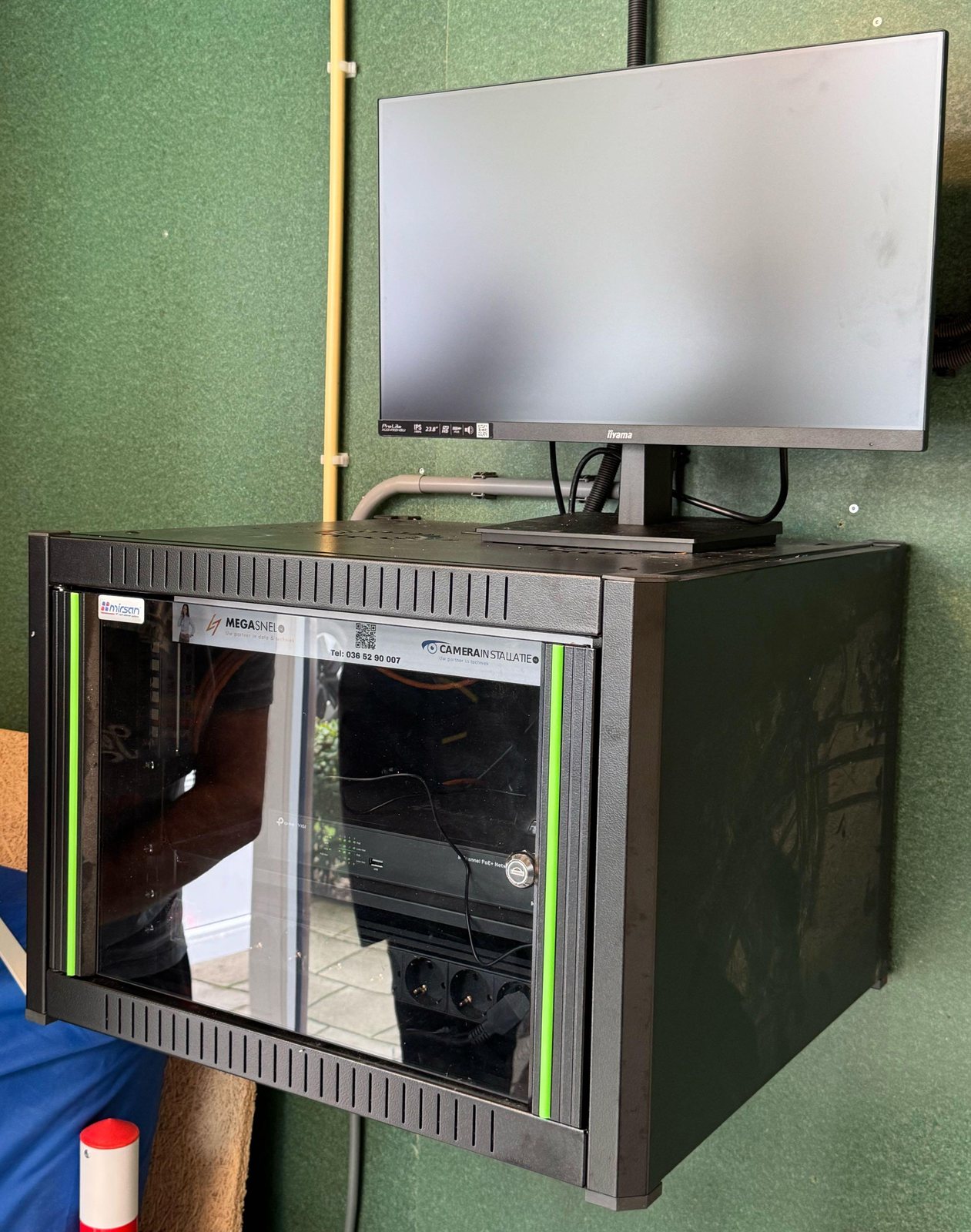 Zwarte wandpatchkast met glazen deur, met daarin de VIGI-recorder en een stekkerdoos en bovenop een monitor, in een afgesloten ruimte