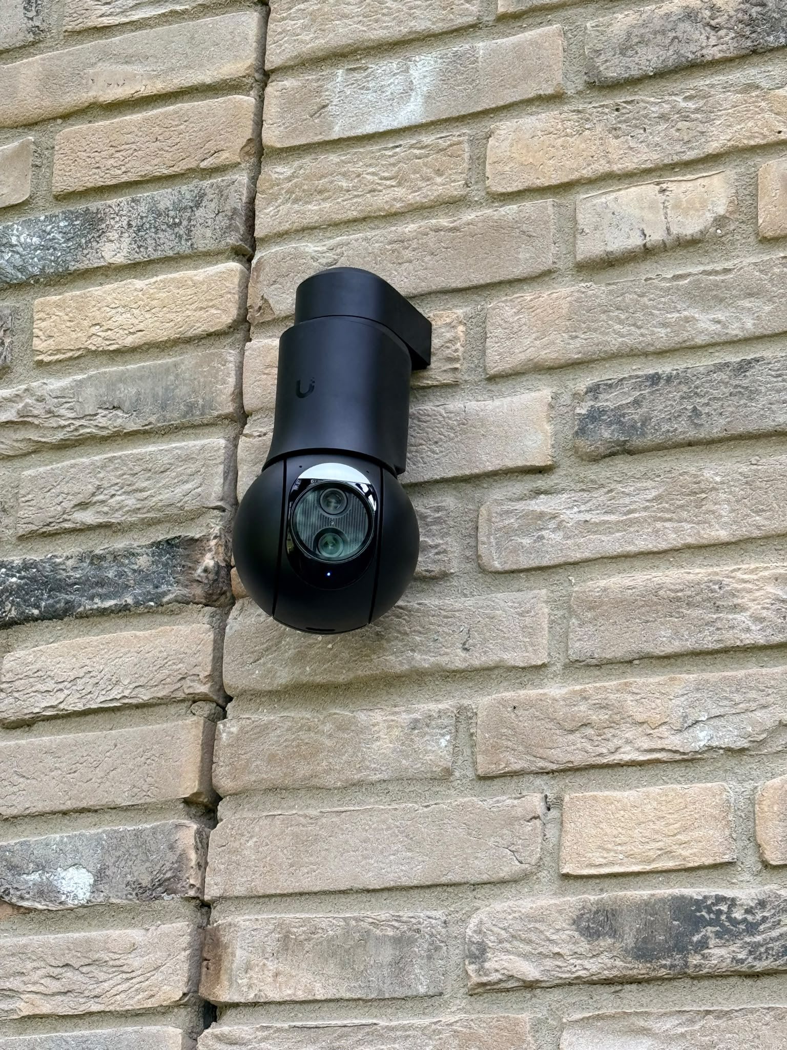 Dichtbij: de zwarte UniFi PTZ-camera met draaibare kop op zijn muurbeugel tegen de baksteen