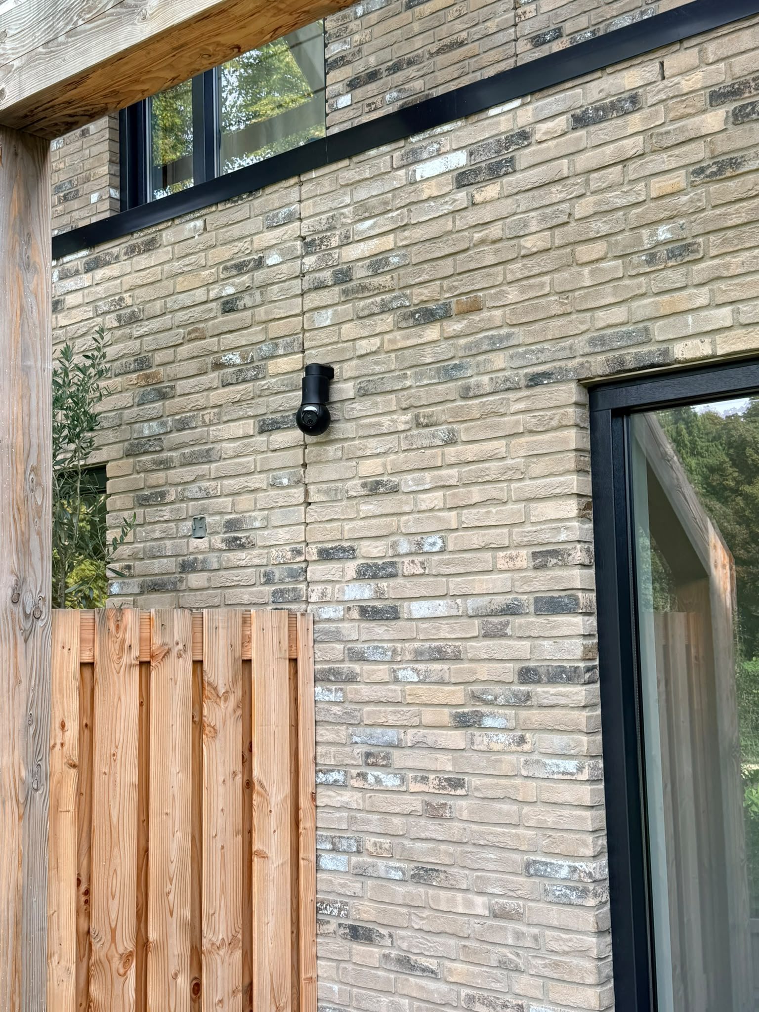 Zwarte UniFi PTZ-camera op de bakstenen achtergevel van een nieuwbouwwoning, boven de houten schutting en naast de glazen tuindeur