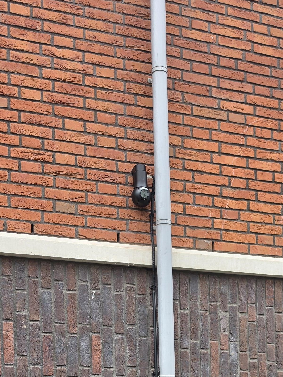 Zwarte UniFi-camera op een muurarm aan de bakstenen gevel, naast een grijze regenpijp