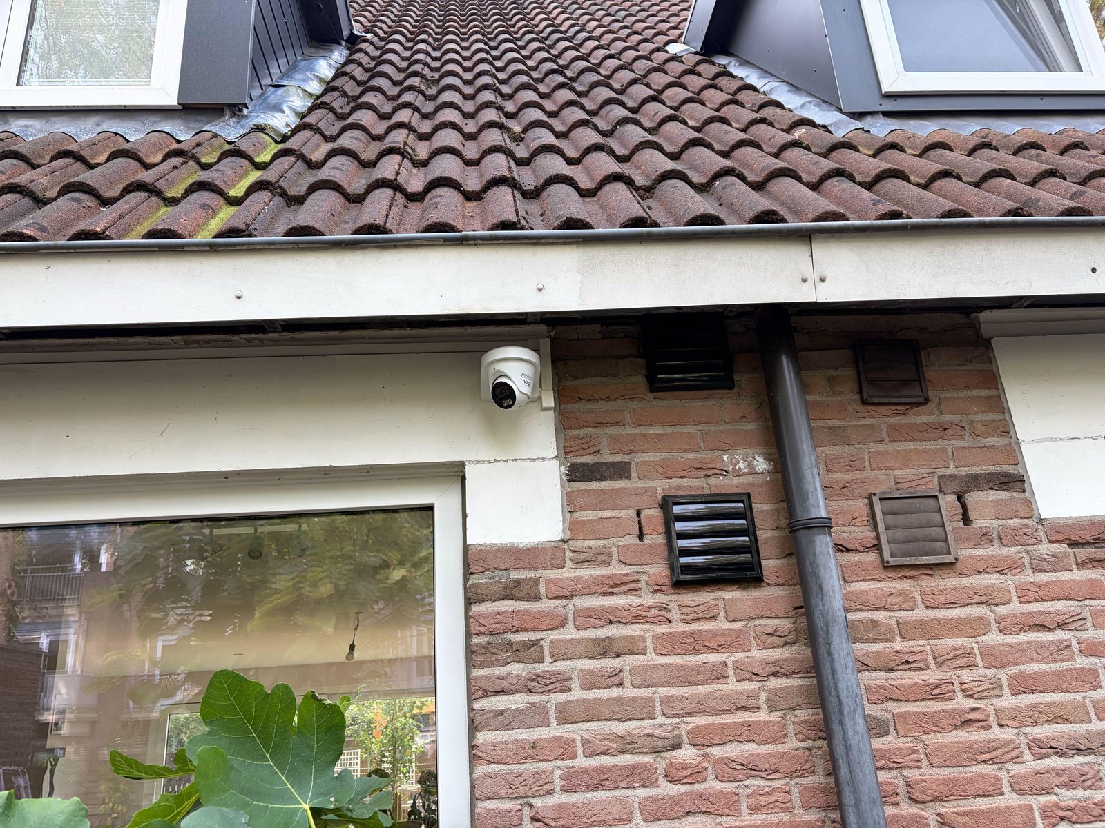 VIGI-camera onder de dakgoot aan de bakstenen achtergevel, naast de regenpijp en boven de tuindeur