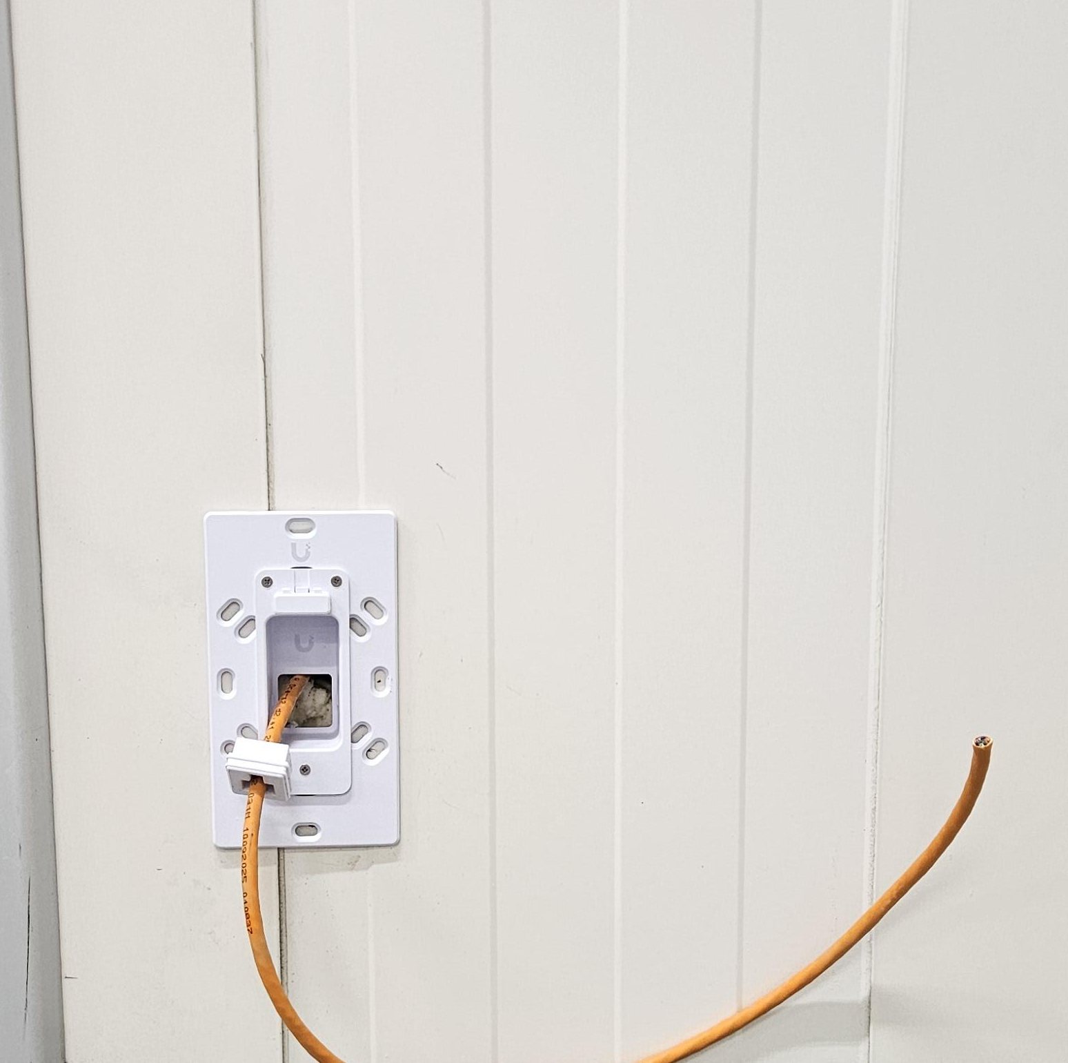 Witte inbouwplaat van een UniFi-lezer op een witte wand, met een oranje netwerkkabel eruit