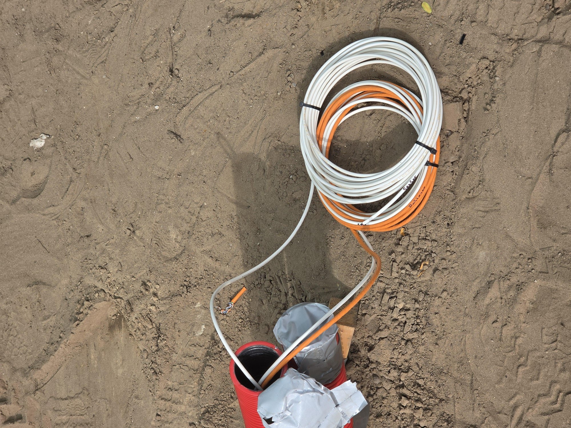 Rollen witte en oranje kabel in het zand bij een rode mantelbuis, tijdens de bouw van de stalling