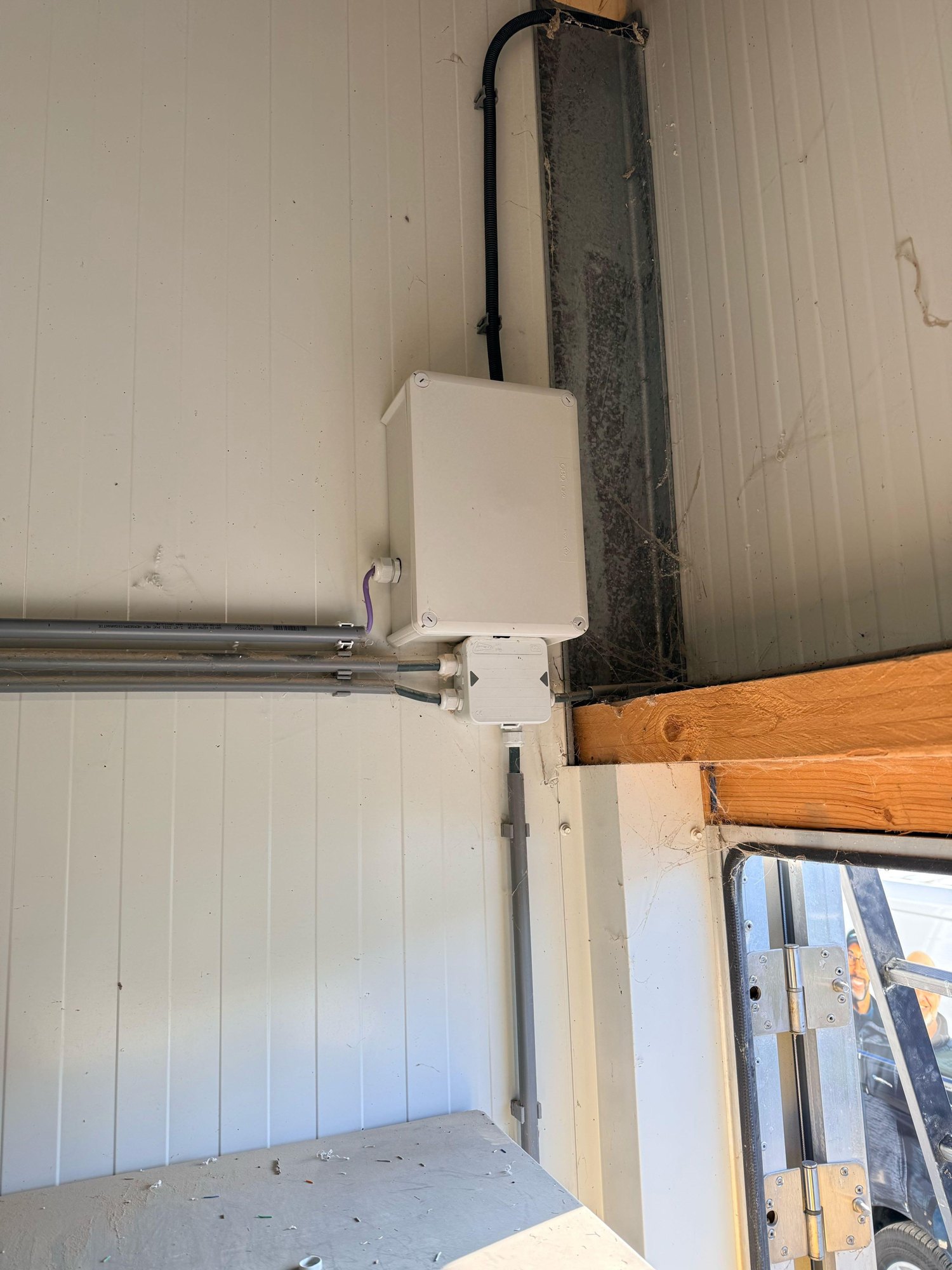 Grijze kast met een lasdoos eronder op een witte stalwand, met buizen en kabel naar de straalzender