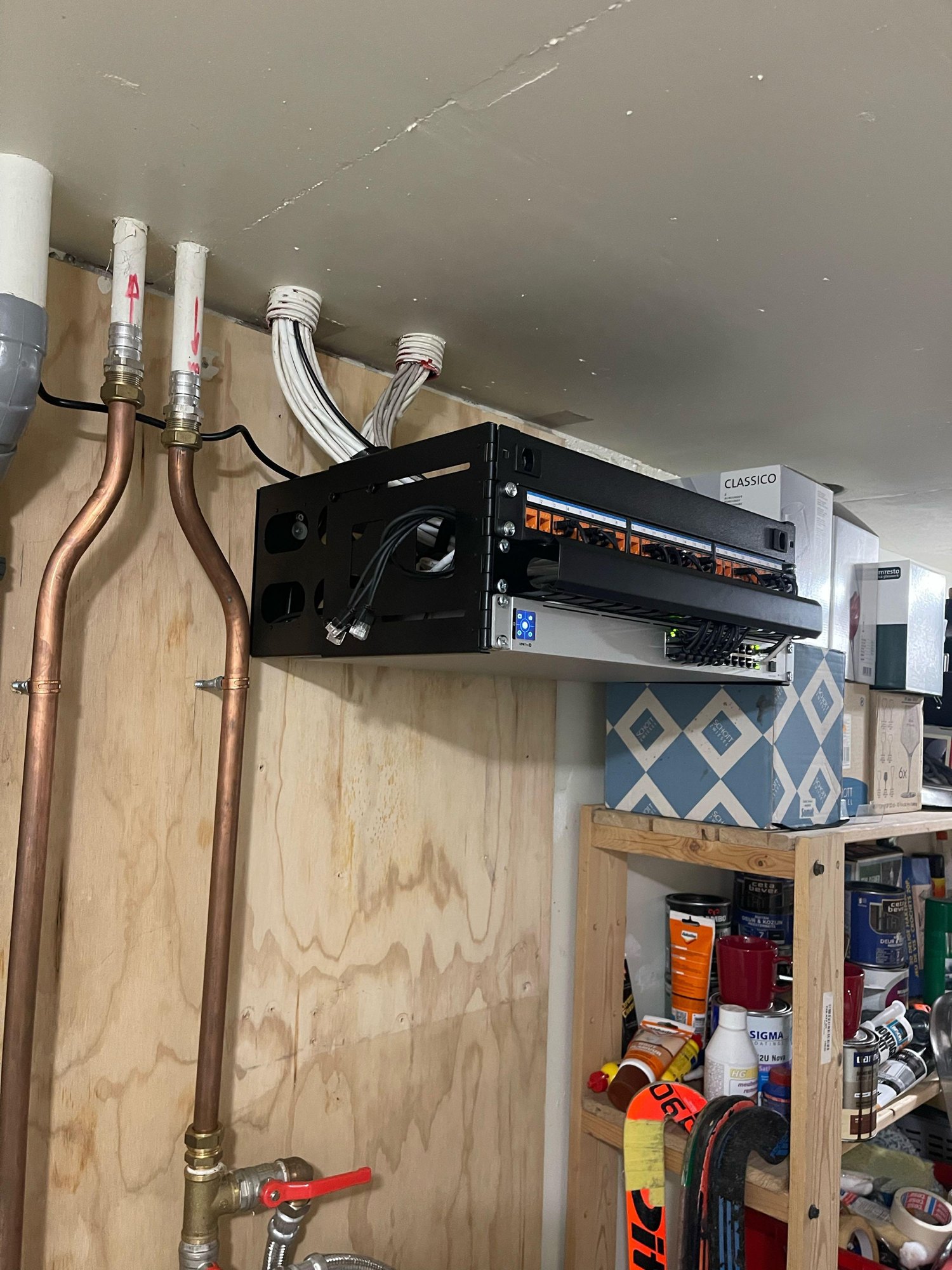 Zwart open patchrek van 4U aan een houten wand, met een rangeerpaneel met oranje keystones en een UniFi-switch eronder, de netwerkkabels netjes vanuit het plafond