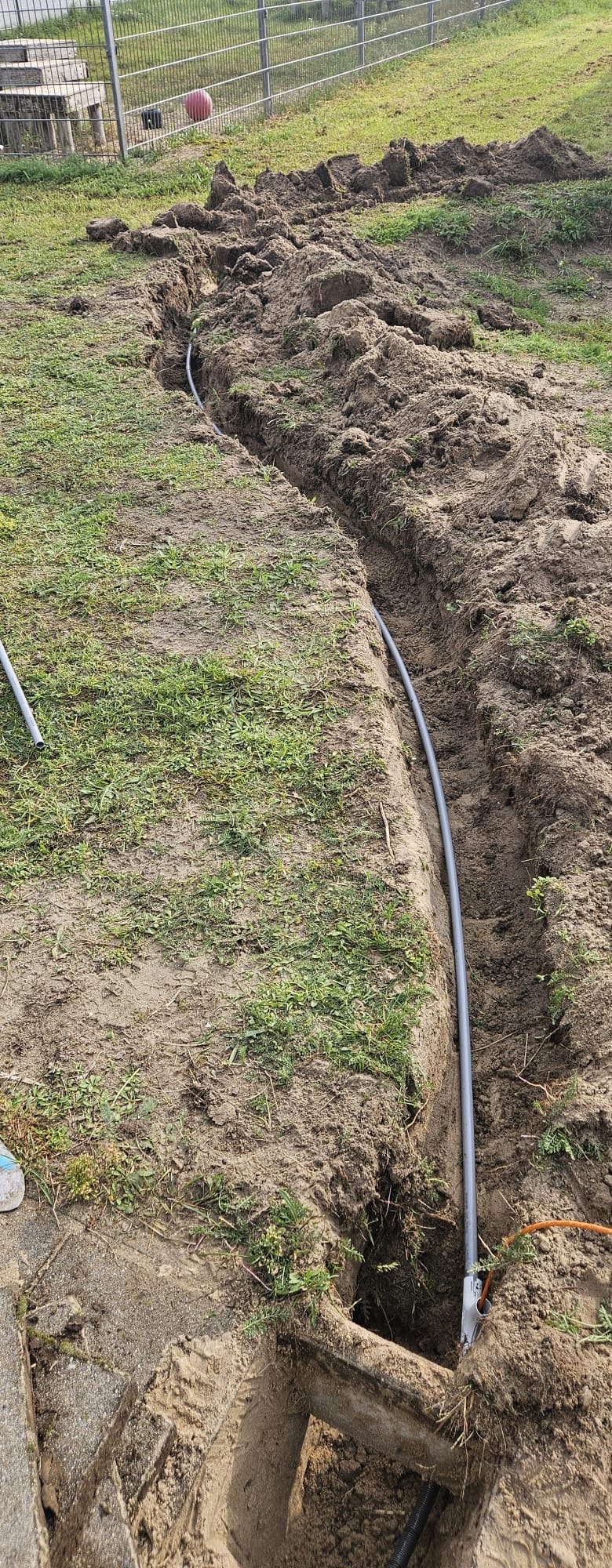 Lange sleuf in het gras met grijze mantelbuis en een oranje kabel, richting een camerapaal