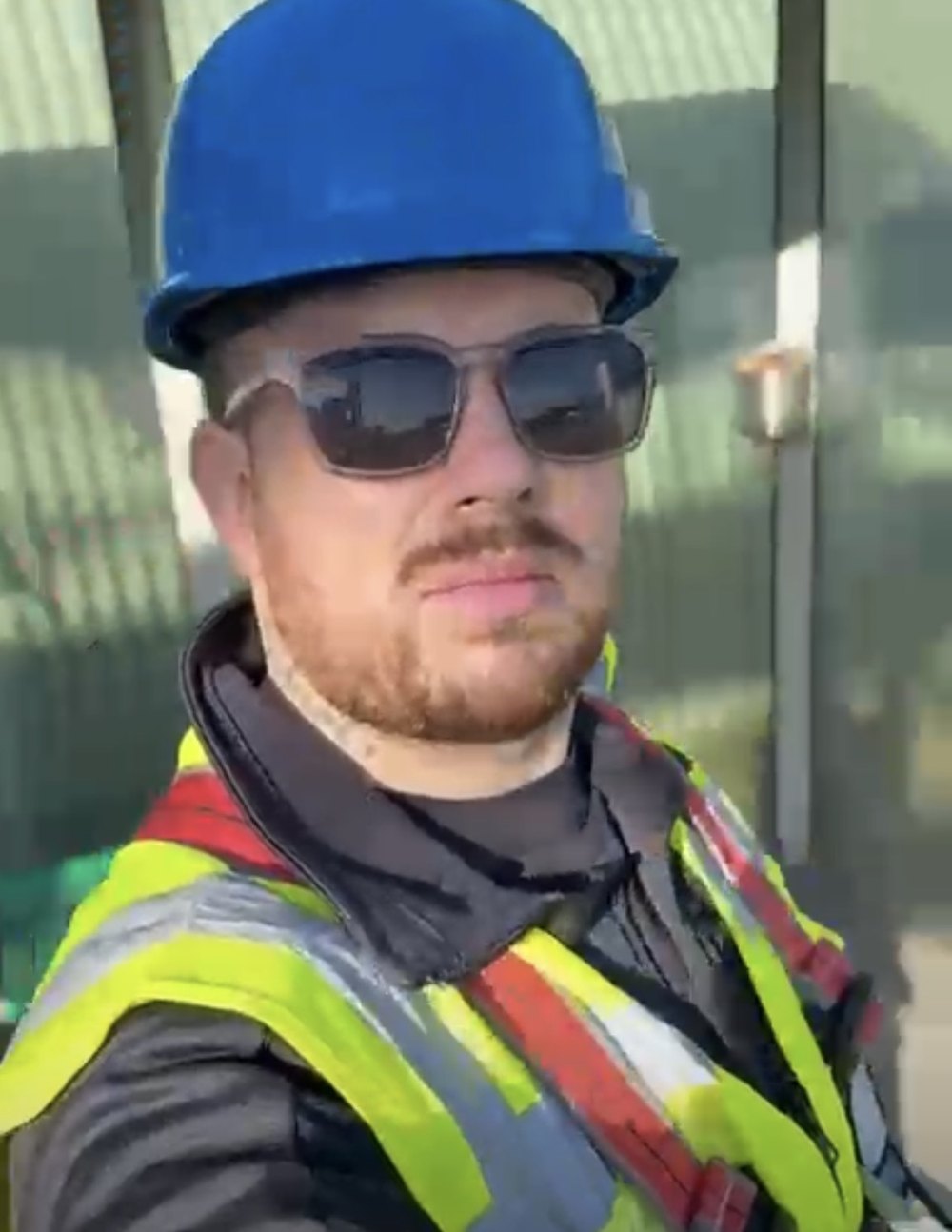 Onze monteur met blauwe helm en veiligheidshesje op het productieterrein, voor een groene afrastering