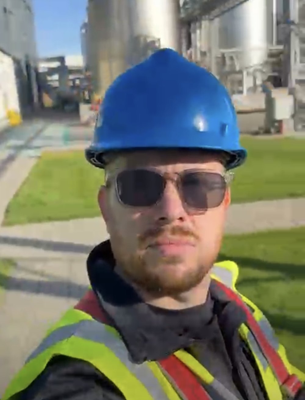 Onze monteur met helm en veiligheidshesje op het productieterrein, met de opslagtanks achter hem